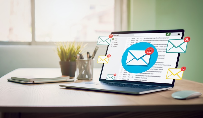 A laptop on a wooden desk displays an email inbox with multiple new email notifications, represented by envelope icons with numbers indicating unread messages. The desk also has a plant, pen holder, and a window in the background allowing natural light to pour over the scene.