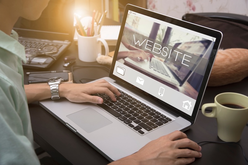 A person using a laptop at a desk, viewing the word "WEBSITE" and several icons, searches how to remove negative content from Google. A notebook, cup of coffee, and phone sit nearby as sunlight streams in from the left.